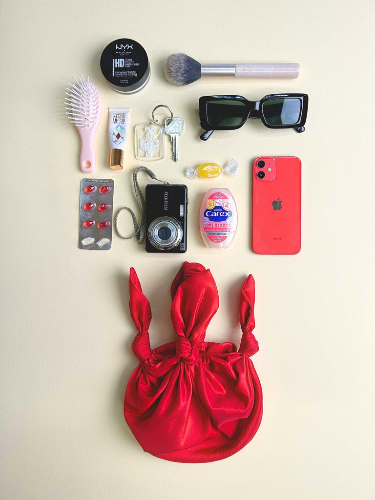 Collection of personal items including a phone, camera, and makeup items on a beige background. A red taffeta handbag sits at the bottom.