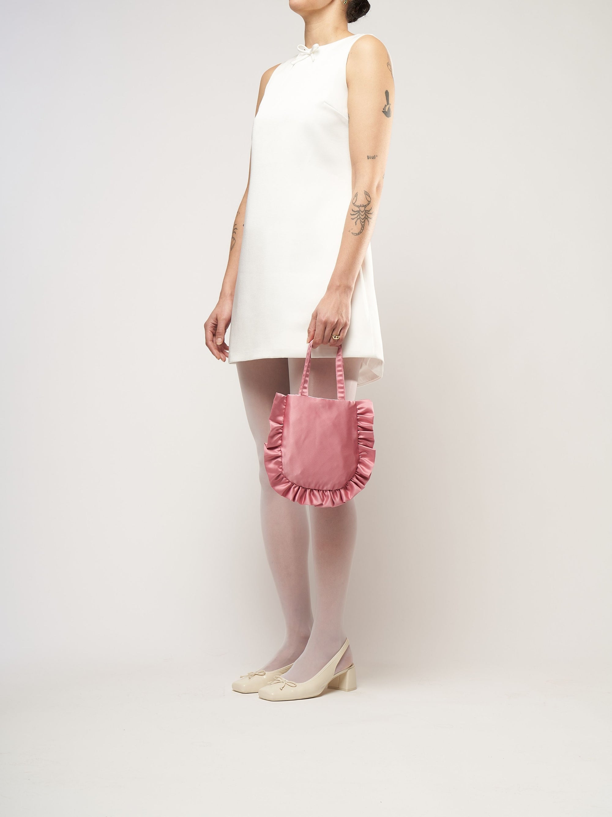 A person in a white sleeveless dress stands against a plain background. They hold a small pink bag and wear white tights with cream heels.