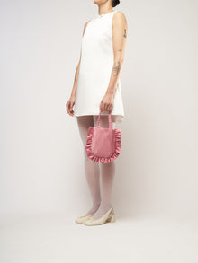 A person in a white sleeveless dress stands against a plain background. They hold a small pink bag and wear white tights with cream heels.