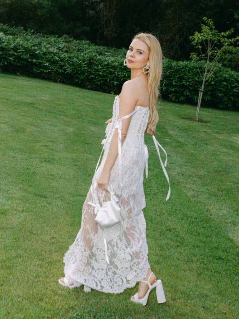 Saoirse-Monica Jackson in a white lace wedding dress holding a small ivory satin ruched handle handbag standing on grass with greenery in the background