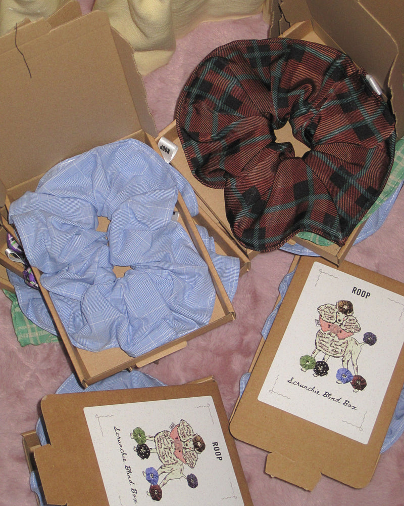 Cardboard boxes on a soft pink rug. The boxes contain large scrunchies, blue plaid, brown tartan and green check. The design on the box is a vintage inspired poodle which scrunchies accessories. They are labelled 'scrunchie blind box'.