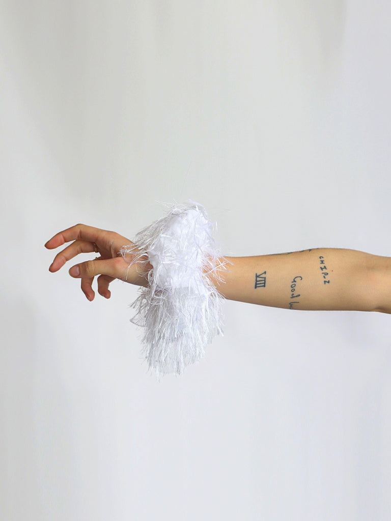 Hand wearing a white feathered scrunchie around their wrist, set against a plain background.