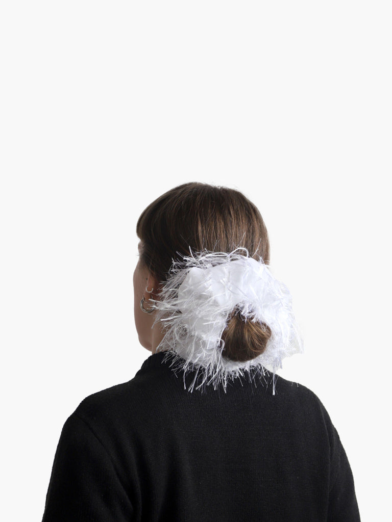 Person wearing a black knit cardigan with a white feathered scrunchie around their bun, set against on a white background.