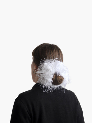 Person wearing a black knit cardigan with a white feathered scrunchie around their bun, set against on a white background.