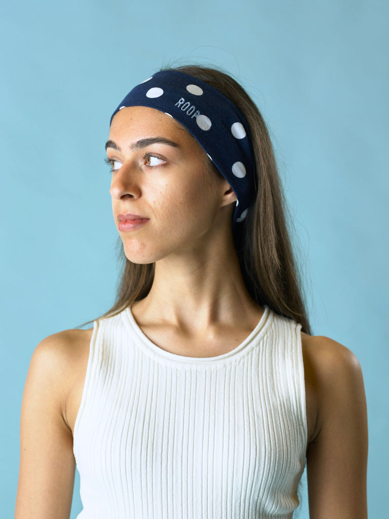 Woman wearing a navy blue stretch headband with white polka dots and ROOP branding against a light blue background