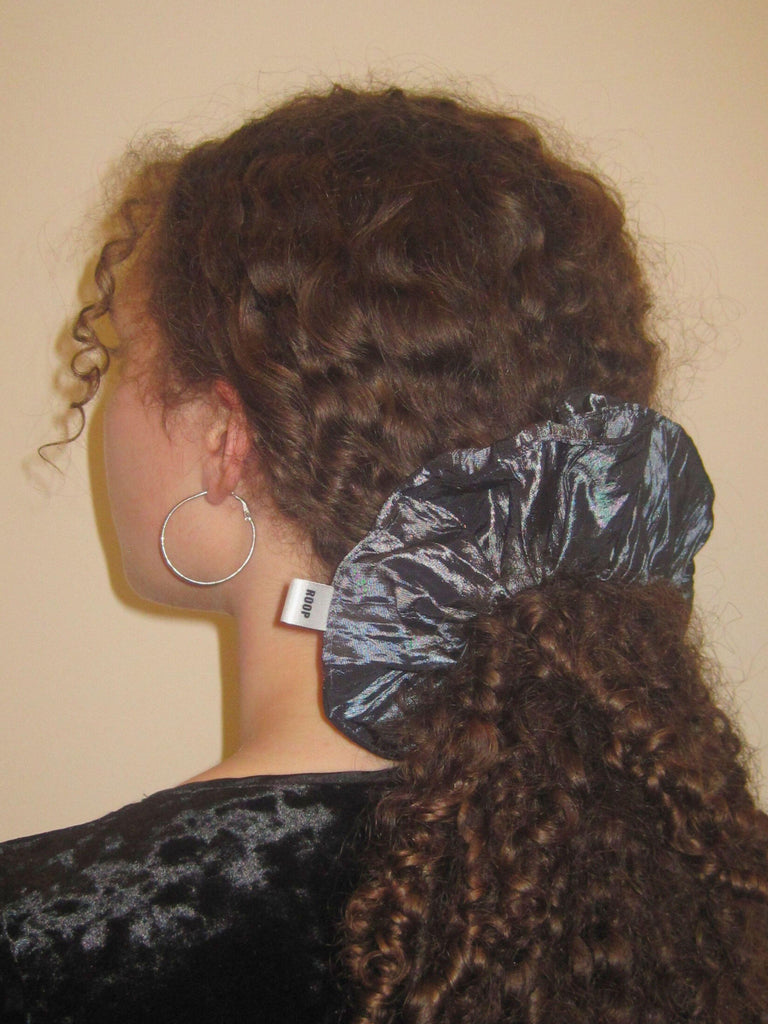 Person with curly brown hair wears a large slate grey scrunchie in a ponytail, they have a black velvet top on and silver hoop earrings, set against on beige background
