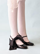 A pair of legs wearing white lace tights and patent black mary jane heels. A black satin hair bow scrunchie is perched on top of one of the shoes. Set in a pale blue background.
