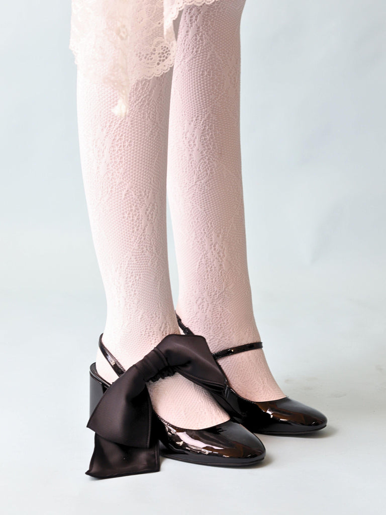 A pair of legs wearing white lace tights and patent black mary jane heels. A black satin hair bow scrunchie is perched on top of one of the shoes. Set in a pale blue background.