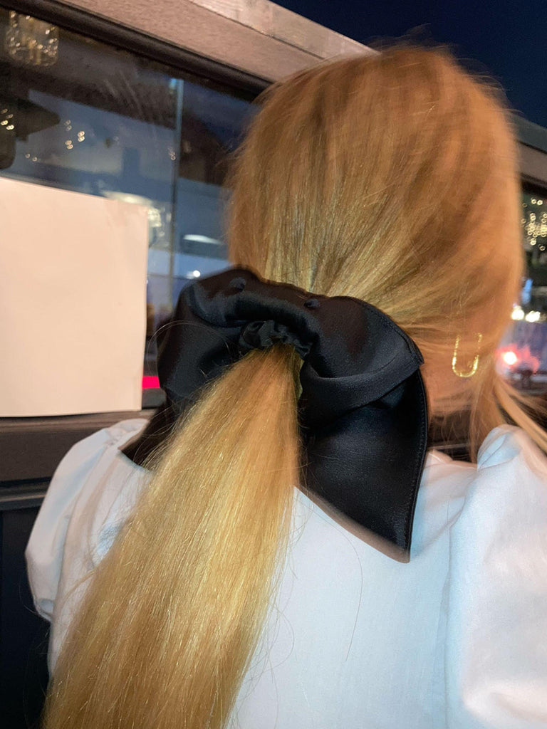 Person with a long blonde ponytail has a black satin hair bow scrunchie in and wears a white puff sleeve top. The background is blurred.