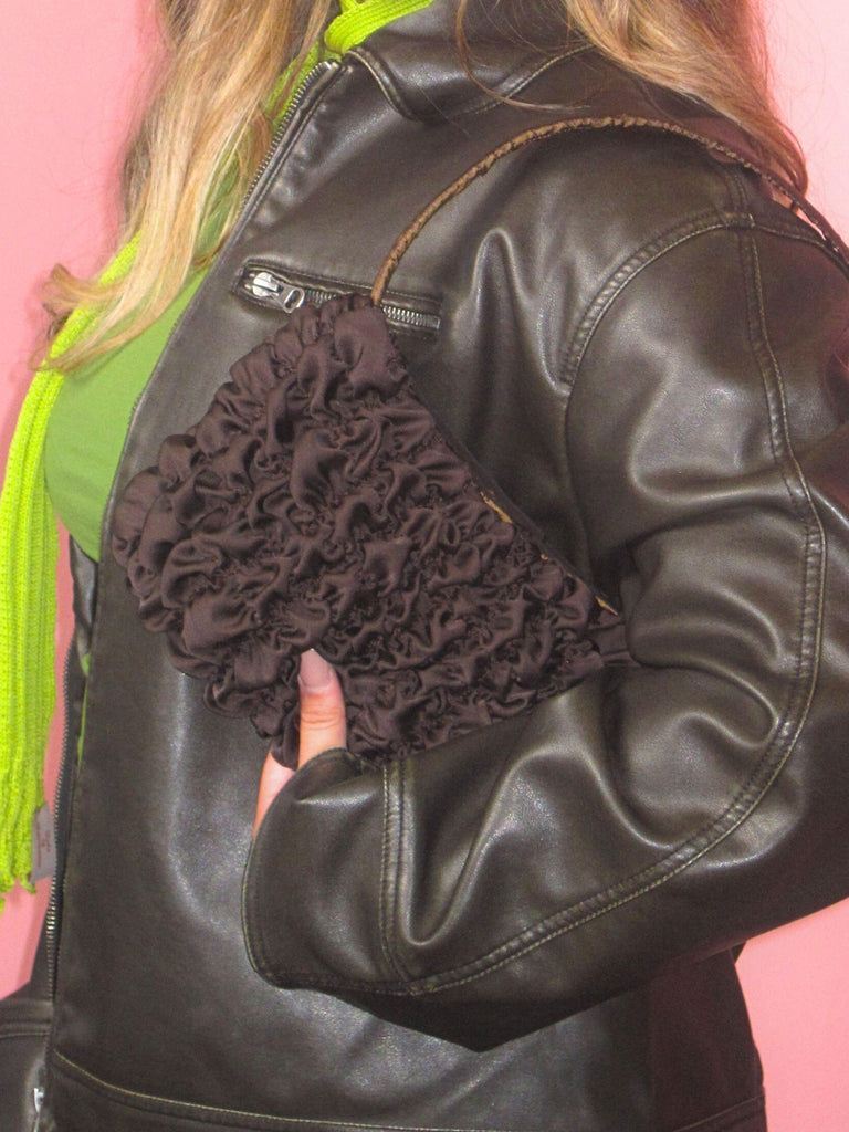 Crop in of a person wearing a brown leather jacket , green top and lime green skinny scary, has a brown textured satin handbag on their shoulder, set against a pink background.
