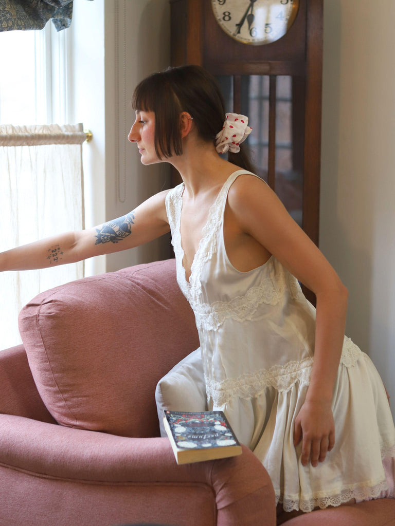 Woman in a white lace nightgown sitting on a pink chair with a book, in a room with a clock and window. She wears a white and pink polka dot scrunchie in her ponytail