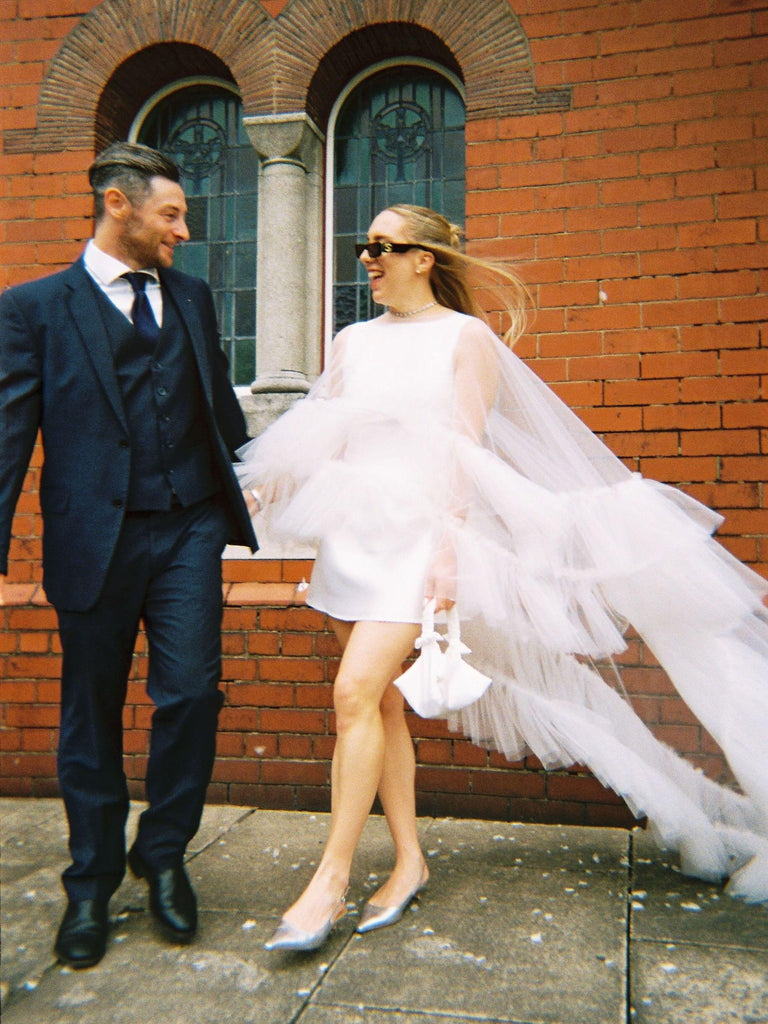 Man and woman in wedding attire standing in front of a brick building. She holds a small ivory satin ruched handle handbag.