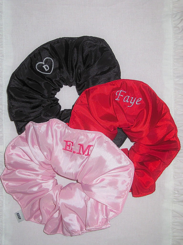 Three taffeta scrunchies in black, red, and pink with embroidered names on a textured white background.