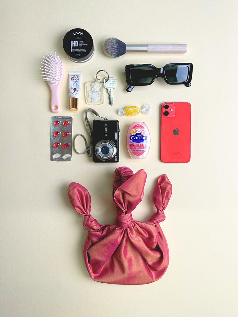 Collection of personal items including a phone, camera, and makeup products on a beige background. pink knotted handbag sits at the bottom.