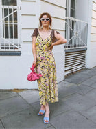 Woman in a floral dress and white sunglasses holds a pink knotted handbag. She is standing on a street with a white building in the background