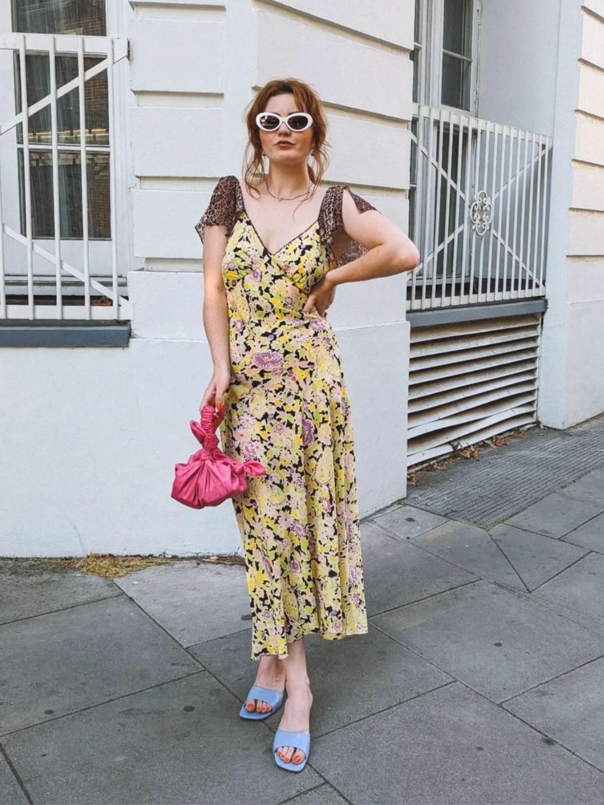 Woman in a floral dress and white sunglasses holds a pink knotted handbag. She is standing on a street with a white building in the background