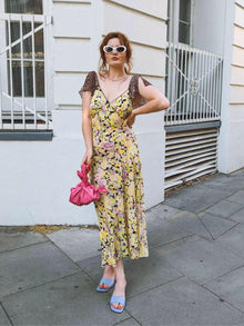 Woman in a floral dress and white sunglasses holds a pink knotted handbag. She is standing on a street with a white building in the background