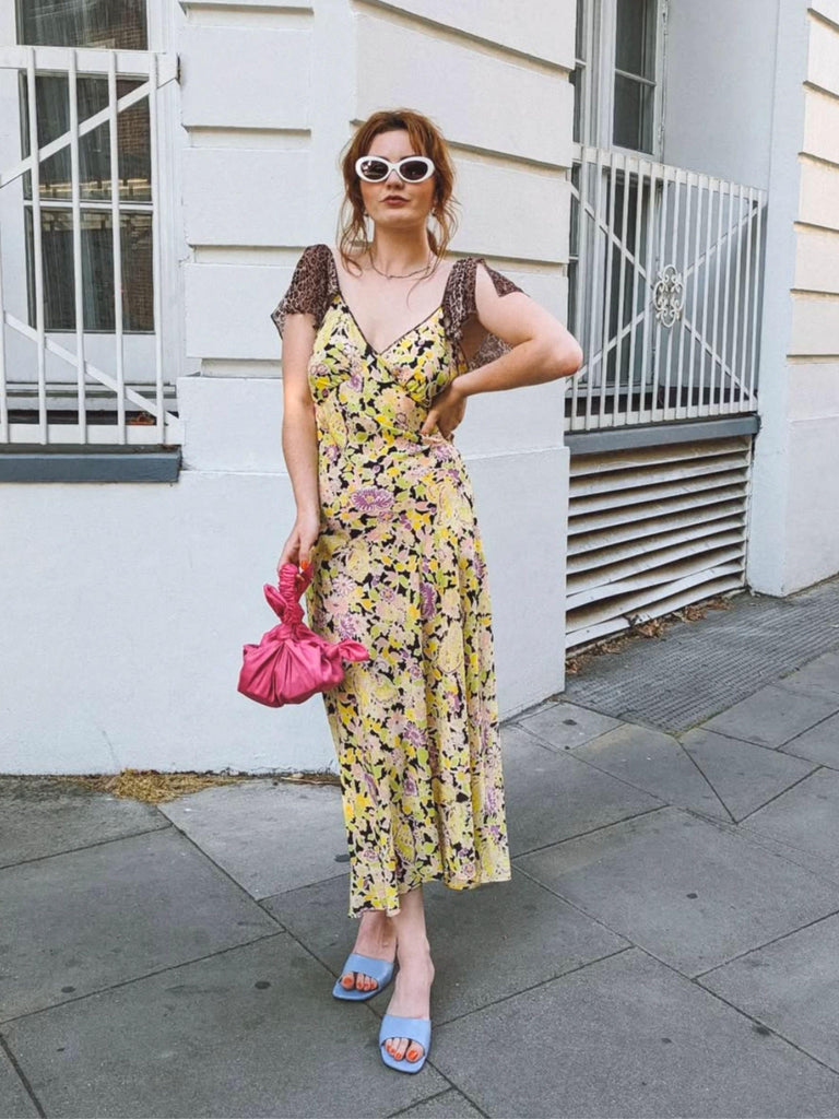 Woman in a floral dress and white sunglasses holds a pink knotted handbag. She is standing on a street with a white building in the background