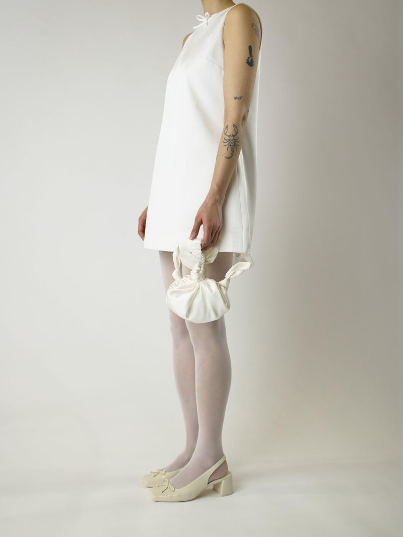 Person wearing an ivory short sleeveless wedding dress, white sjeer tights and heeled ivory ballet shoes, holds a knotted ivory handbag, they are standing against a plain background.