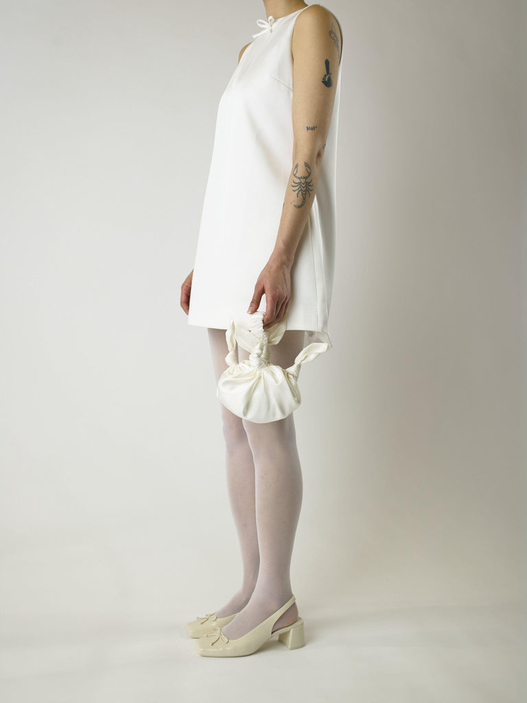 Person wearing an ivory short sleeveless wedding dress, white sjeer tights and heeled ivory ballet shoes, holds a knotted ivory handbag, they are standing against a plain background.