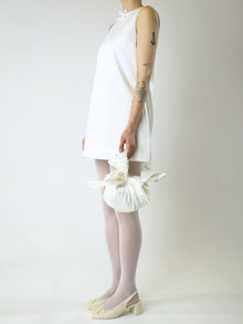 Person wearing an ivory short wedding dress holding an ivory knotted bag against a plain background