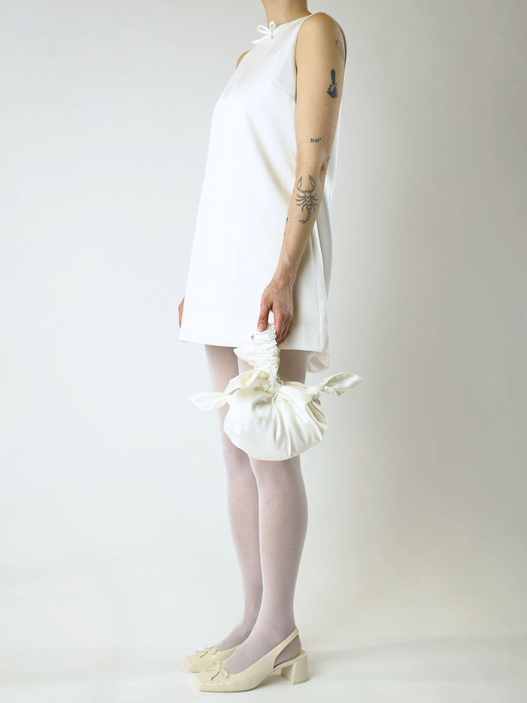 Person wearing an ivory short wedding dress holding an ivory knotted bag against a plain background