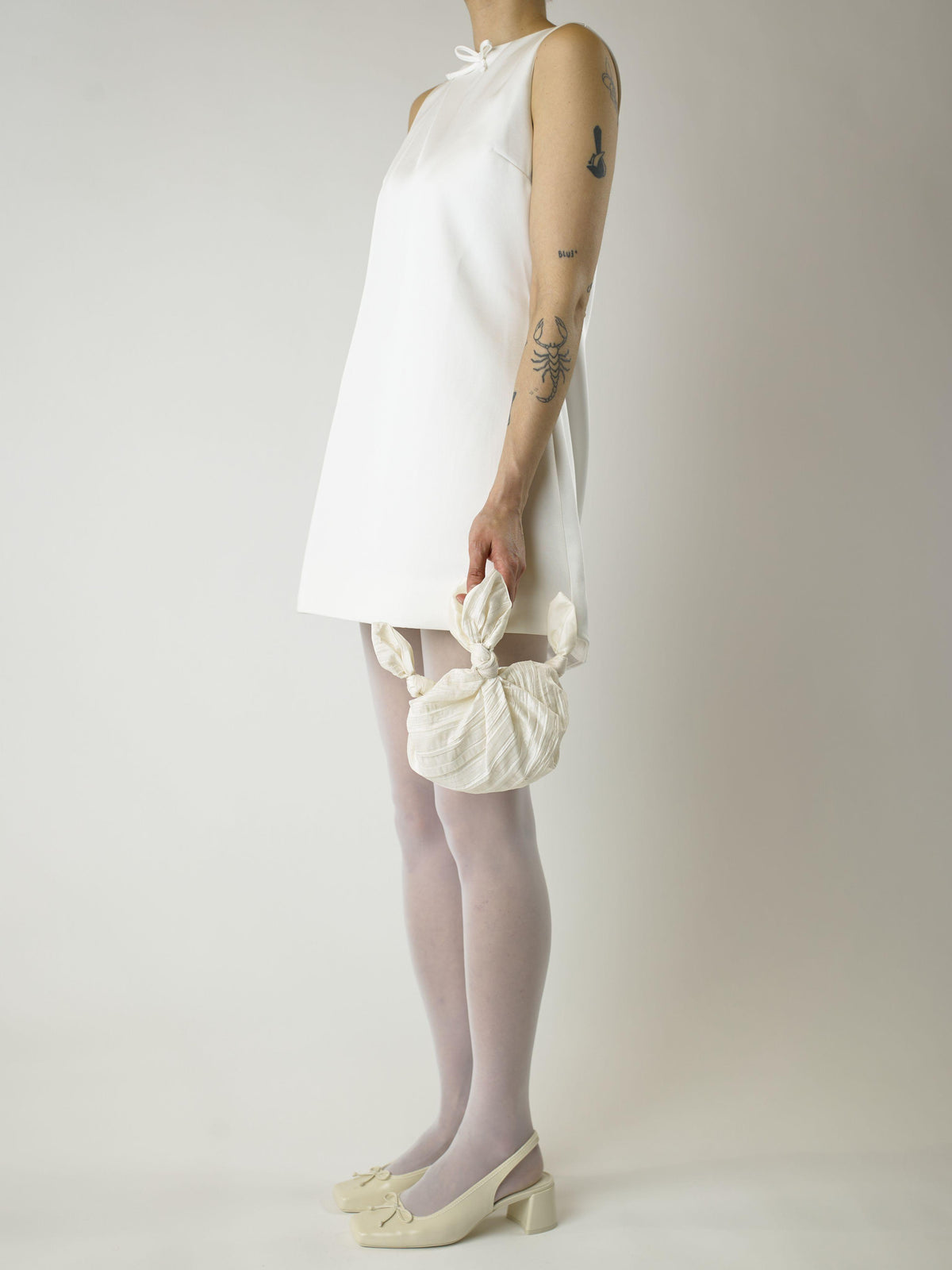 model wearing a short ivory wedding dress and heeled ivory ballet shoes. They're holding an ivory crinkle taffeta bridal handbag. Set on blank background.