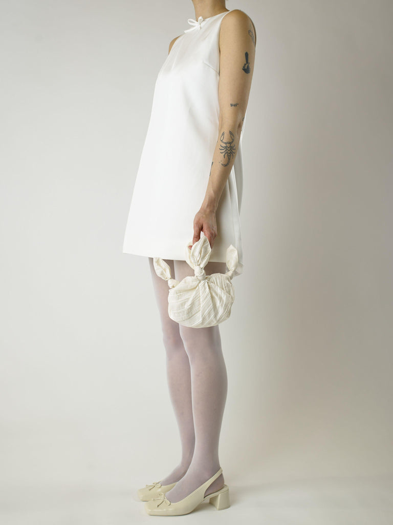 model wearing a short ivory wedding dress and heeled ivory ballet shoes. They're holding an ivory crinkle taffeta bridal handbag. Set on blank background.