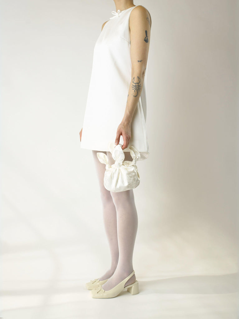 Person wearing an ivory sleeveless short wedding dress and ivory heeled ballet shoes, holds a small ivory knotted satin handbag. They're stood on a plain background