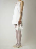 Person wearing an ivory sleeveless wedding dress, white sheer tights and heeled ballet pumps, holding a small ivory satin handbag against a plain background 