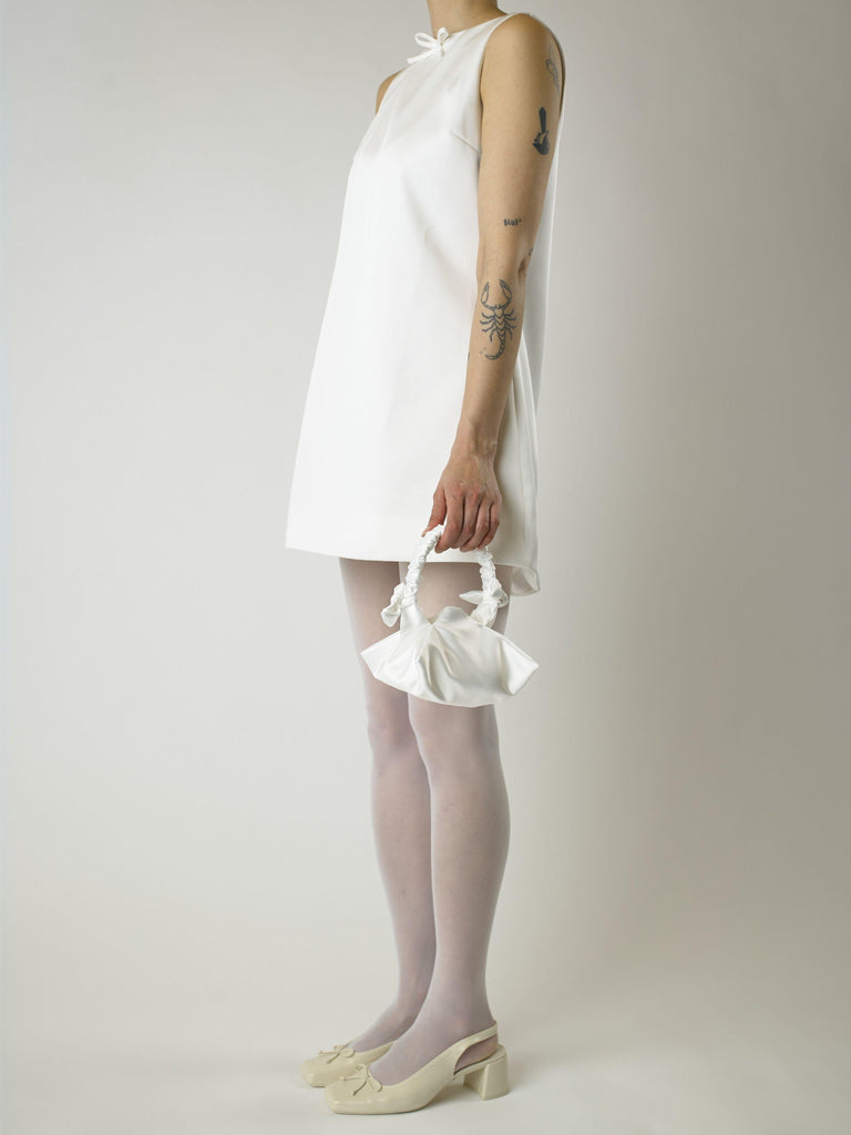 Person wearing an ivory sleeveless wedding dress, white sheer tights and heeled ballet pumps, holding a small ivory satin handbag against a plain background 