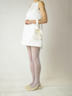Person wearing a short ivory sleeveless wedding dress, sheer stockings and heeled ivory ballet pumps, holds an ivory satin box handbag  with a long handle and bow details on their arm, set against a plain background