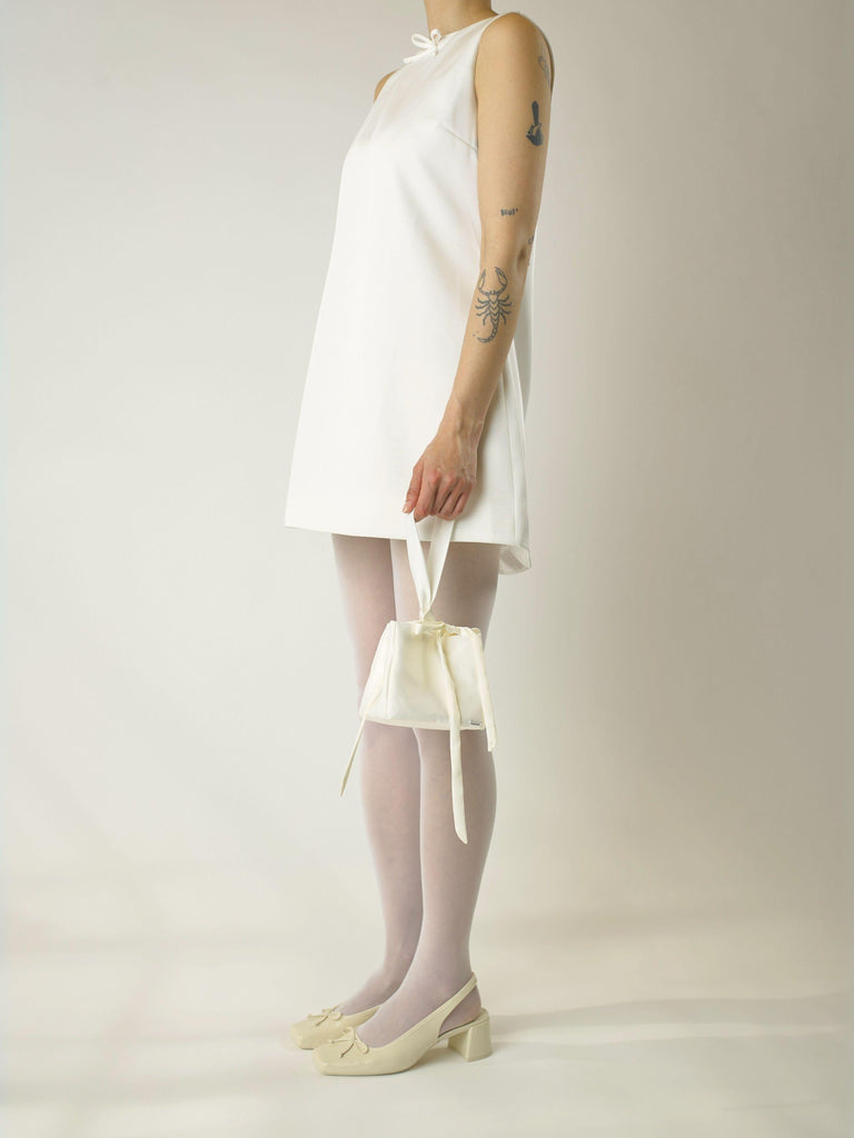 Person wearing a short ivory sleeveless wedding dress, sheer stockings and heeled ivory ballet pumps, holds an ivory satin box handbag with a long handle and bow details, set against a plain background