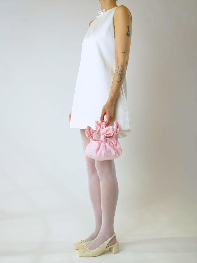 Person wearing a short ivory wedding dress and ivory heeled ballet shoes, holds a pink knotted handbag on a plain background