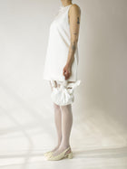Person wearing an ivory short sleeveless wedding dress, heeled ivory ballet shoes and sheer white stocking, holds a white knotted handbag against a plain background