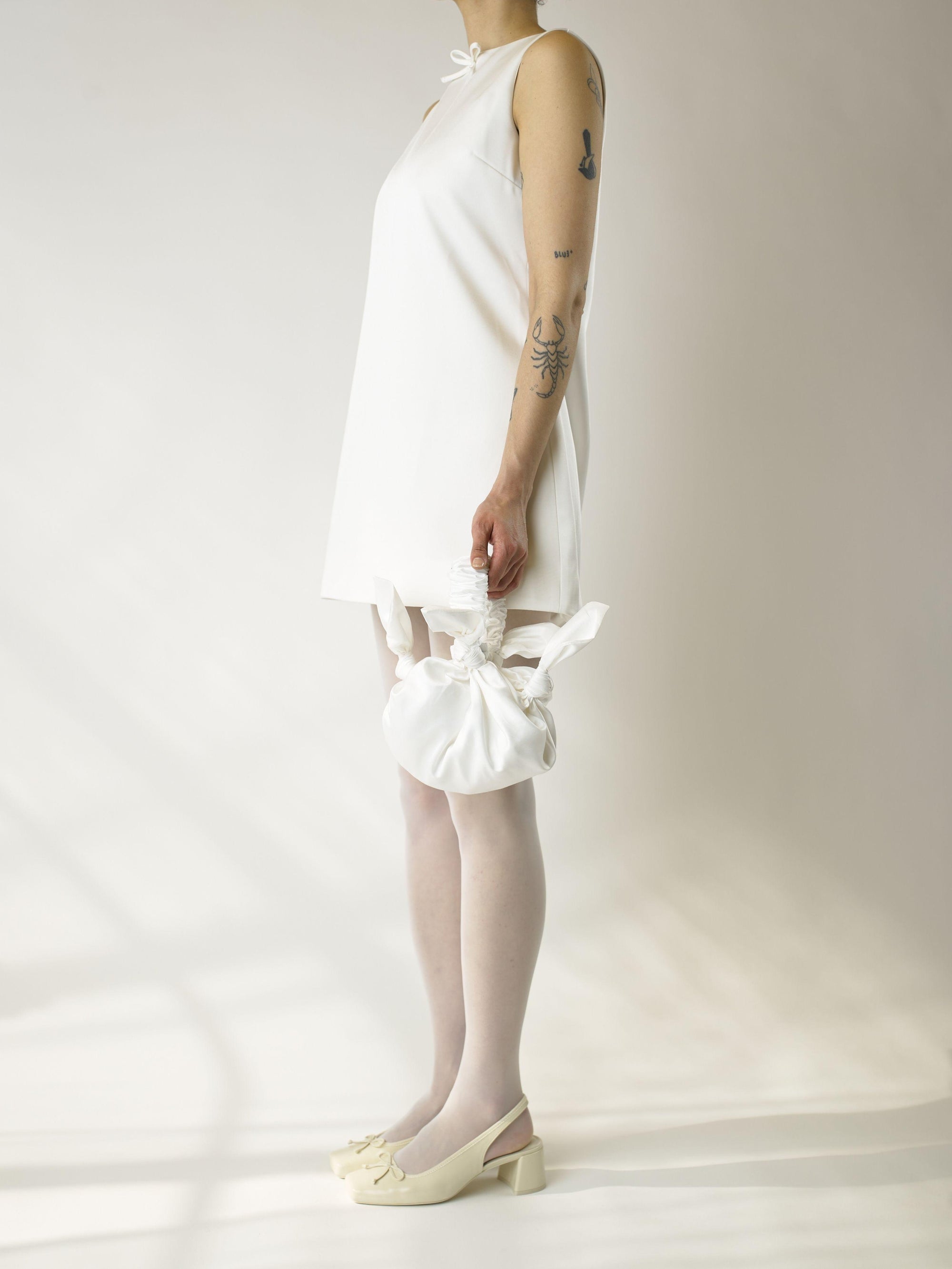 Person wearing an ivory short sleeveless wedding dress, heeled ivory ballet shoes and sheer white stocking, holds a white knotted handbag against a plain background