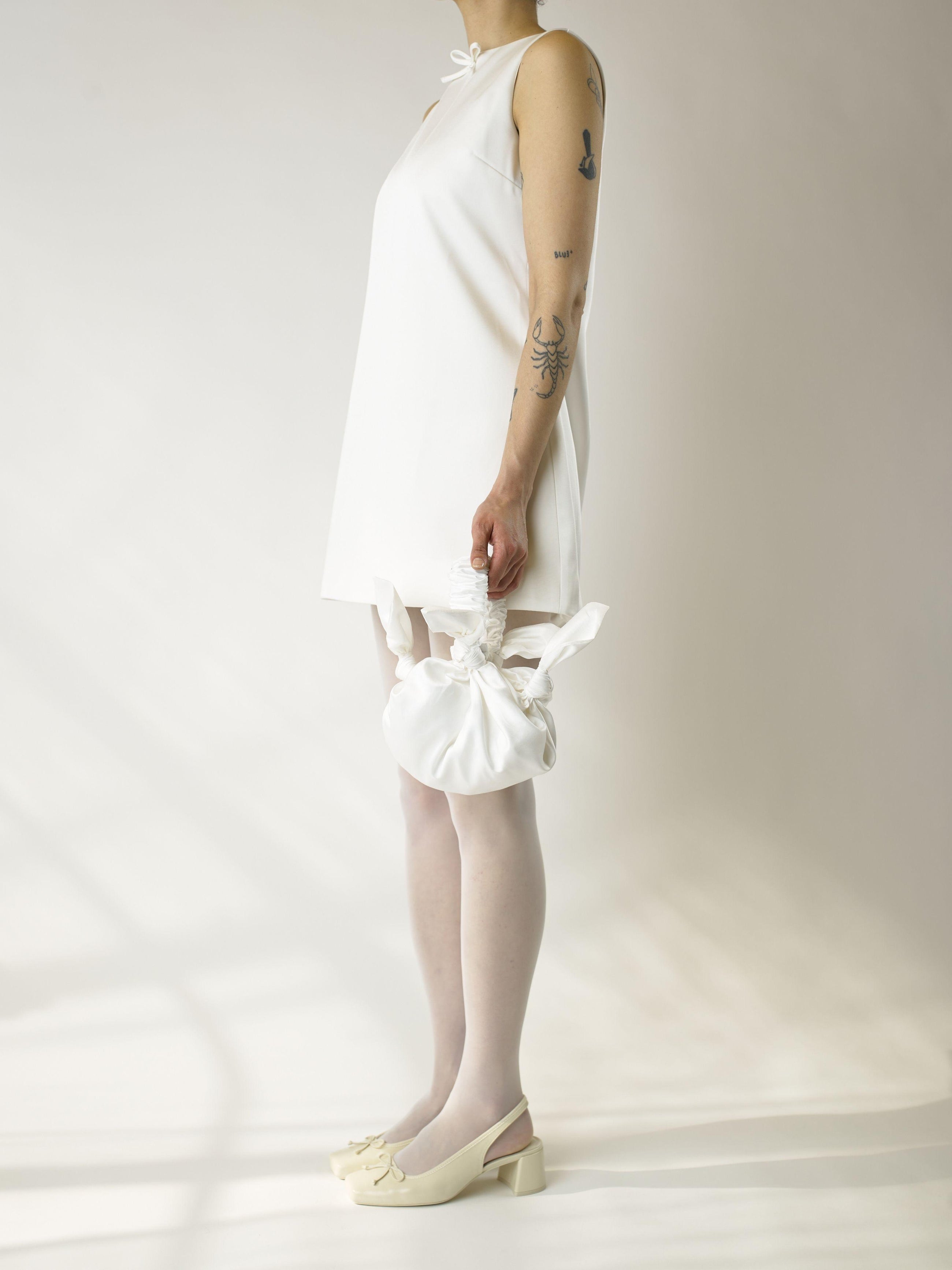 Person wearing an ivory short sleeveless wedding dress, heeled ivory ballet shoes and sheer white stocking, holds a white knotted handbag against a plain background