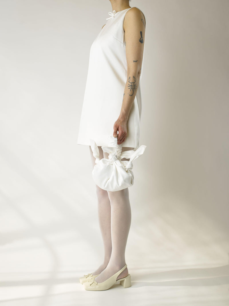 Person wearing an ivory short sleeveless wedding dress, heeled ivory ballet shoes and sheer white stocking, holds a white knotted handbag against a plain background