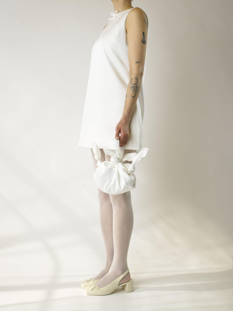 Person wearing an ivory short sleeveless wedding dress, heeled ivory ballet shoes and sheer white stocking, holds a white knotted handbag against a plain background
