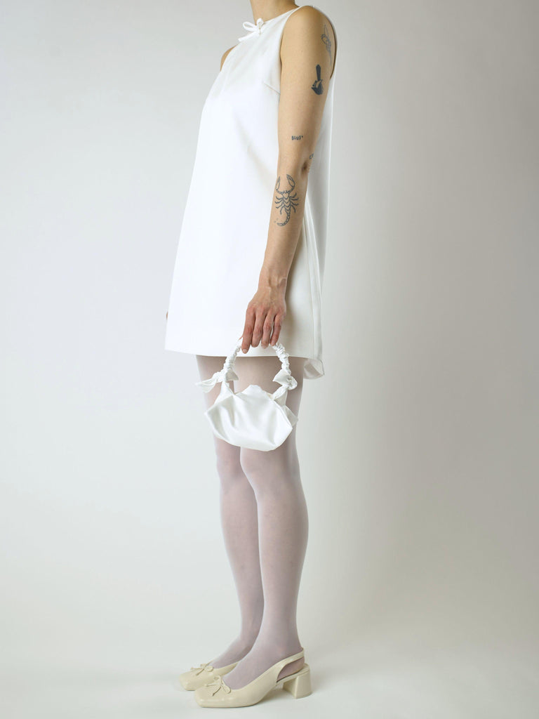 Person wearing an ivory sleeveless wedding dress, white sheer tights and heeled ballet pumps, holding a small white satin handbag against a plain background 