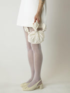 Cropped in image of a model wearing a short ivory wedding dress and heeled ivory ballet shoes. They're holding an ivory crinkle taffeta bridal handbag. Set on blank background.