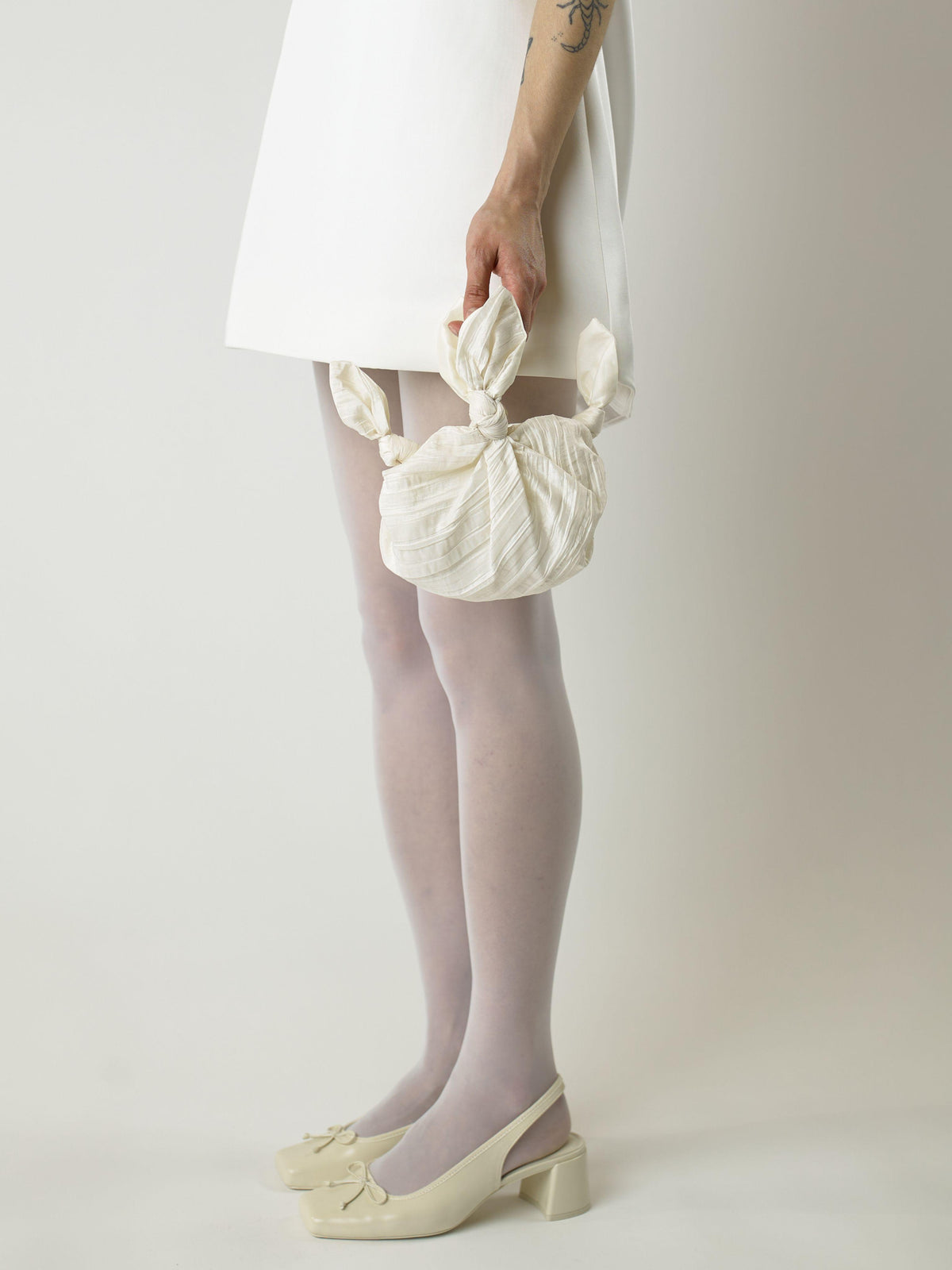 Cropped in image of a model wearing a short ivory wedding dress and heeled ivory ballet shoes. They're holding an ivory crinkle taffeta bridal handbag. Set on blank background.