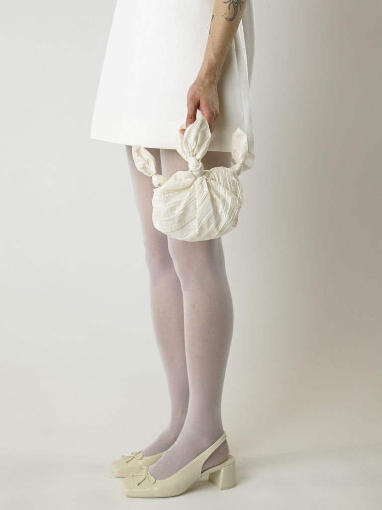 Cropped in image of a model wearing a short ivory wedding dress and heeled ivory ballet shoes. They're holding an ivory crinkle taffeta bridal handbag. Set on blank background.