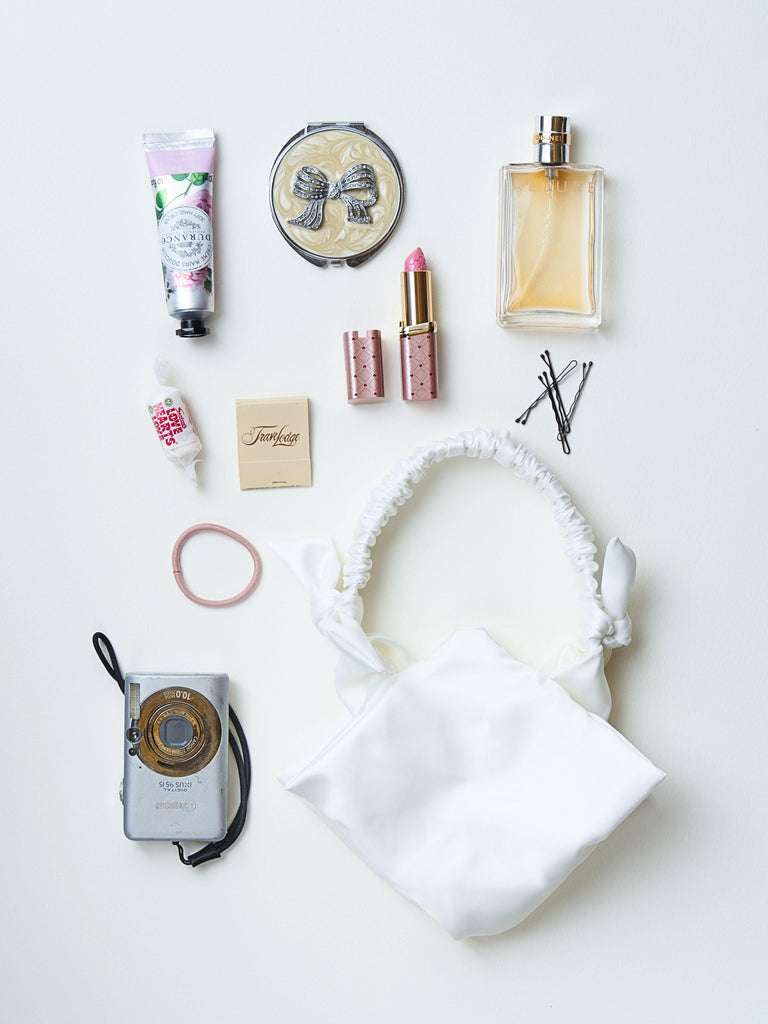 Collection of beauty products including a camera, hair accessories, and skincare items next to a ivory satin ruched handle handbag, set on a white background