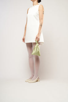 A person in a white sleeveless dress stands against a plain background. They hold a small light green bag and wear white tights with cream heels.