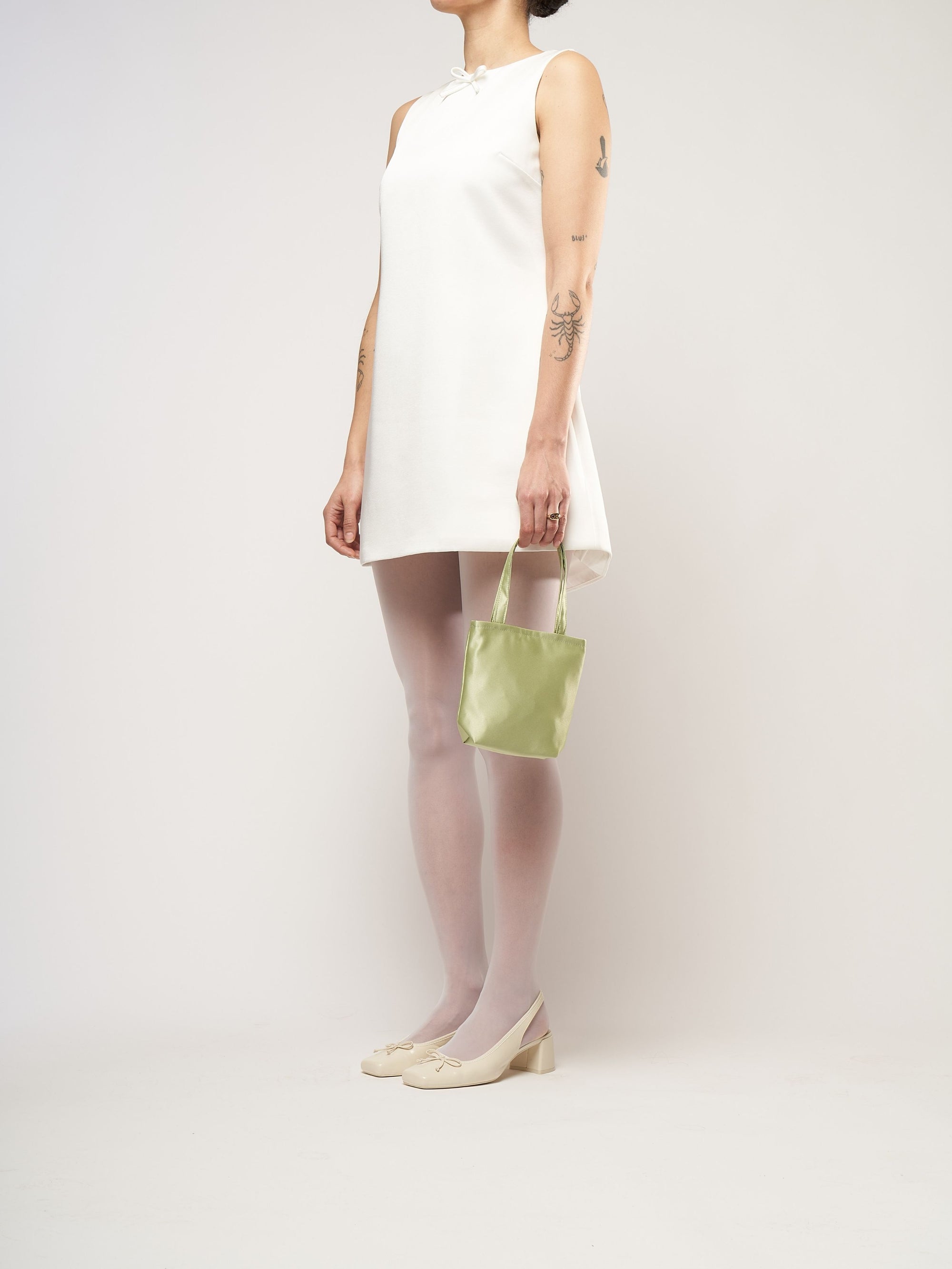 A person in a white sleeveless dress stands against a plain background. They hold a small light green bag and wear white tights with cream heels.