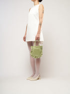 A person in a white sleeveless dress stands against a plain background. They hold a small light green bag and wear white tights with cream heels.