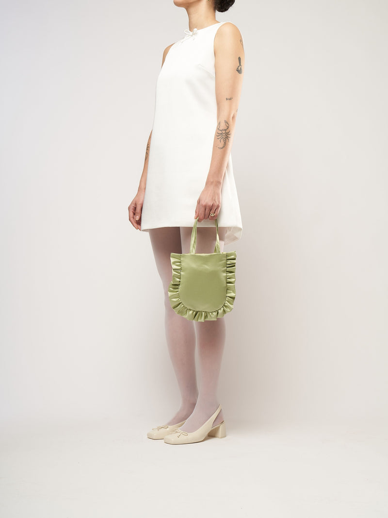 A person in a white sleeveless dress stands against a plain background. They hold a small light green bag and wear white tights with cream heels.