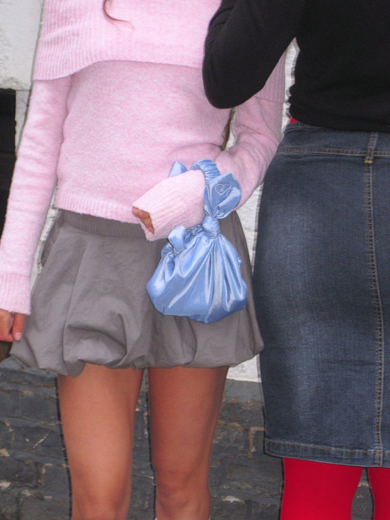 Two people wearing skirts with a focus on their legs and lower bodies. One wears a pink fluffy jumper and holds a lilac knotted handbag.