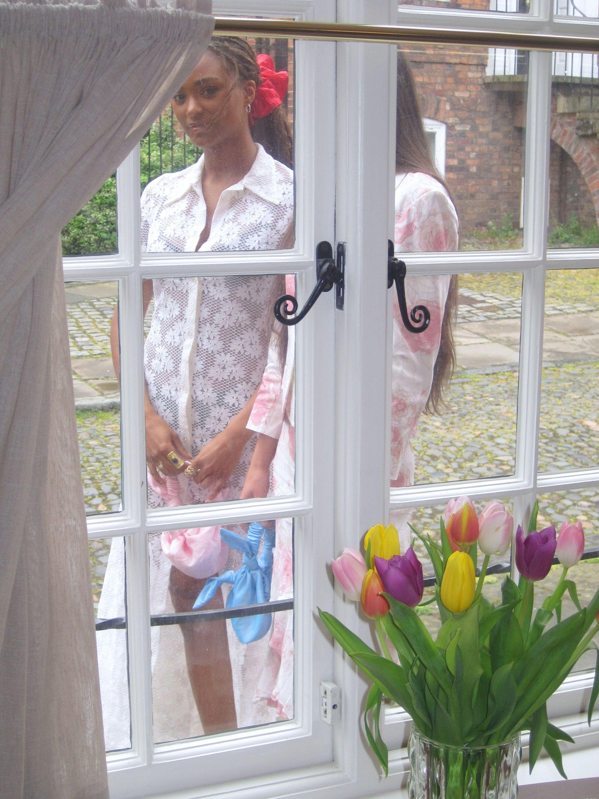 Woman standing behind a window with a vase of flowers in the foreground. They hold small knotted handbags and one wears a large pink scrunchie.